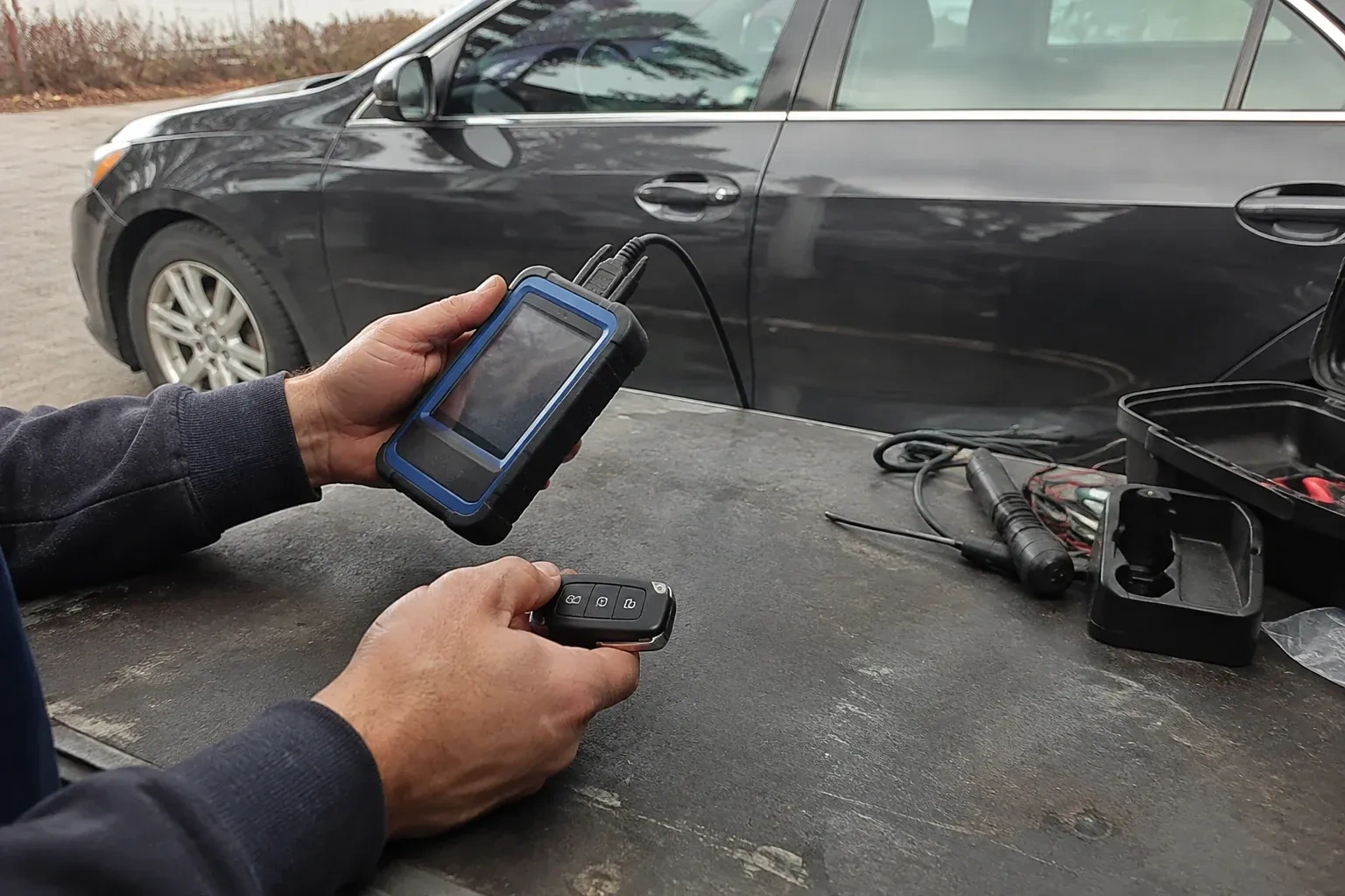 Hands holding a blue-edged electronic device and car-key fob at a worktable beside a parked car. — Transponder Key Programming, Toronto